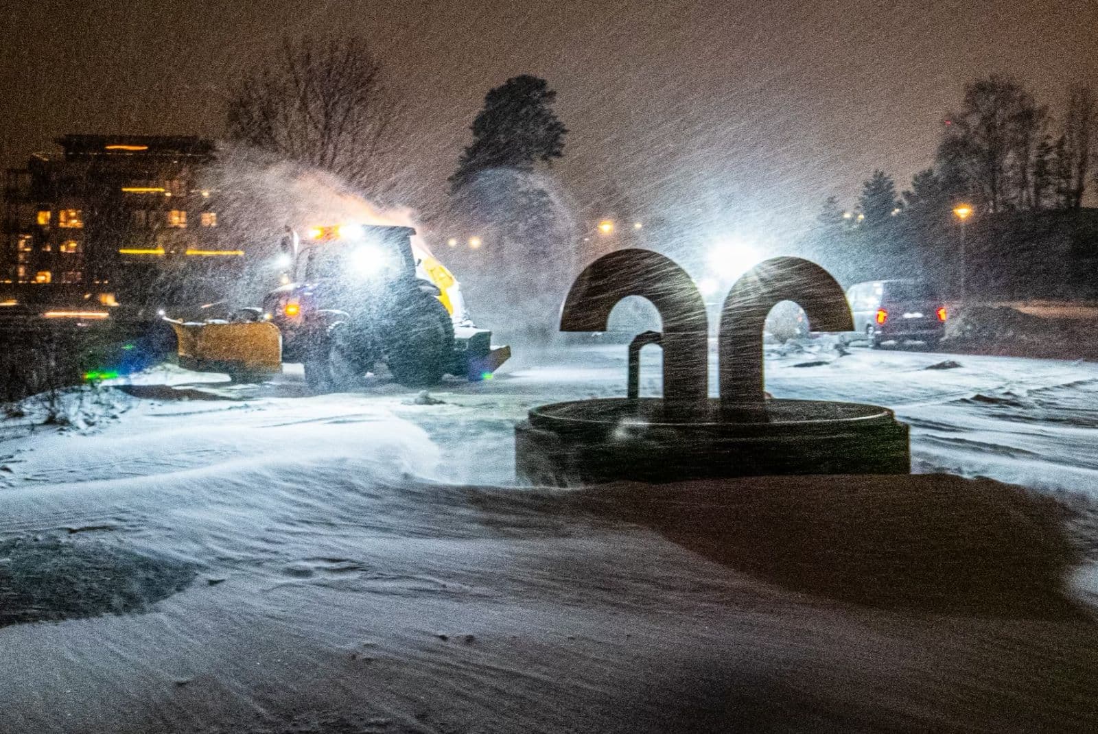 Traktor brøyter en stor plass i snøvær om kvelden