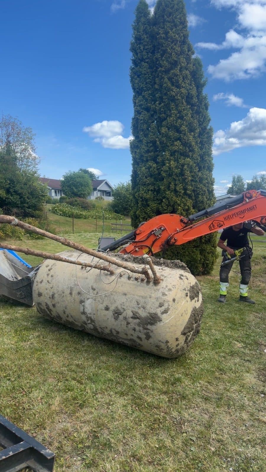 Gravemaskin løfter en gammel, jordete oljetank opp av bakken i en hage